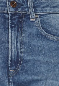 Jeans de mezclilla azul con una textura ligeramente deslavada, presentando un hardware de botón en bronce, costuras en tono marrón y un diseño de cinco bolsillos.