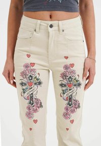 Beige denimjeans med blommiga och korsdesigns i rosa, rött och blått, med hjärtan och logodetaljer på båda benen.