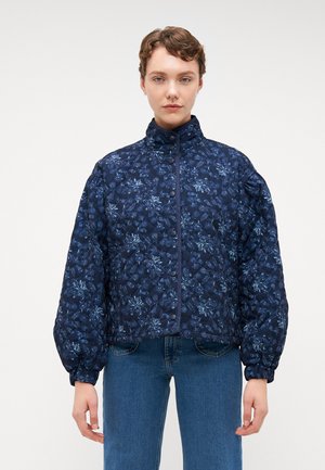 Marineblå quiltet jakke med blomster mønster, høy krage og knappe lukking. Har puffede ermer og elastiske mansjetter. Sammen med blå jeans.