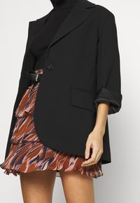 Blazer negro con un solo botón, corte oversized y puños doblados, combinado con una falda con volantes que presenta patrones abstractos en tonos marrón y rosa.