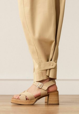 Beige heeled sandal with ankle buckle strap worn with beige cuffed pants on wooden floor.
