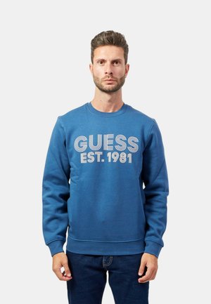 Felpa blu con collo tondo, maniche lunghe e una grafica frontale con "GUESS EST. 1981" in bianco, realizzata in misto cotone.