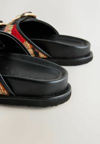 Sandali neri con suole spesse e cinturini decorati con perline multicolori in tonalità di rosso, arancione e oro, su uno sfondo chiaro.