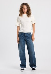 Light beige fitted T-shirt with short, puffed sleeves, paired with loose blue denim jeans and black-and-white sneakers. Simple design, no patterns.
