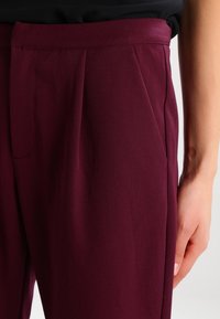 Pantalons bourgogne avec une texture lisse, dotés de plis avant et d'une coupe ajustée. La main repose sur le côté, mettant en valeur le détail de la taille.