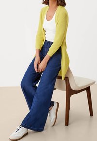 Dlouhý pletený cardigan v jasně žluté barvě, nošený přes bílý top bez rukávů. Doplněno širokými tmavě modrými džíny a bílými teniskami s hnědými podrážkami.