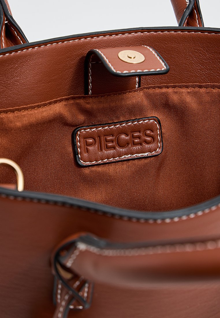 Intérieur d'un sac à main en cuir marron avec une doublure satinée, une étiquette en relief portant l'inscription "PIECES" et une fermeture à pression magnétique.