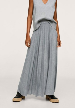 Kobieta ubrana w szary bezrękawnik z dzianiny, długą plisowaną szarą spódnicę i czarne botki z beżowymi grubymi podeszwami stojąca na jednolitym tle.