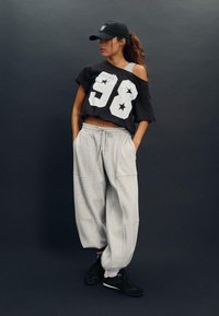 Crna oversized majica s bijelim otiskom "98" i zvijezdama, uparena sa svijetlosivim trenirkama. Outfit je dodatno upotpunjen crnim tenisicama i kapom.