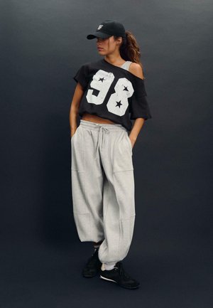 Črna oversize majica s belim printom "98" in zvezdic, v kombinaciji s svetlo sivo trenirko. Outfit dopolnjujejo črne superge in kapa.