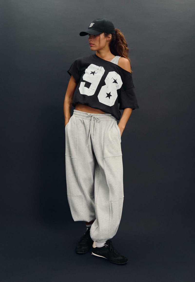 Crna oversized majica s bijelim otiskom "98" i zvijezdama, uparena sa svijetlosivim trenirkama. Outfit je dodatno upotpunjen crnim tenisicama i kapom.