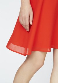 Rotes Kleid aus transparenter Stoffart, mit einem ausgestellten Saum, glatter Textur und einem dezenten Lagen-Effekt am unteren Ende. Eine Hand ist teilweise sichtbar.