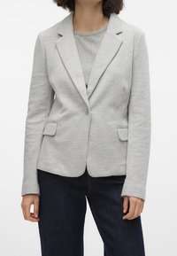 Grijze blazer van katoenmix met inkepingen op de revers, twee zakken aan de voorkant en een enkele knoopsluiting, gedragen over een grijze top en donkere spijkerbroek.