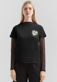 Schwarzes Baumwoll-T-Shirt mit Rundhalsausschnitt, kurzen Ärmeln und einem grafischen Druck auf der Brust. Darunter wird ein langärmliges, transparentes Leo-Print-Oberteil getragen.