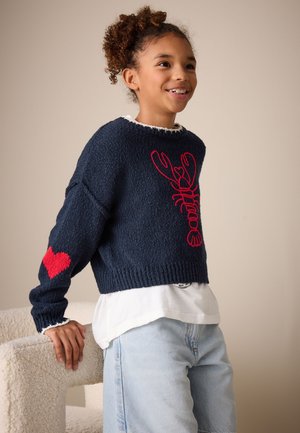 Niño con cabello rizado que lleva un suéter azul marino con diseño de langosta roja y corazón rojo en la manga, apoyado en una silla blanca texturizada.