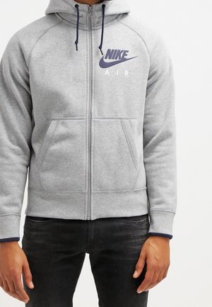 Persona che indossa una felpa con cappuccio grigio chiaro con zip, tasche frontali e logo Nike Air blu navy sul petto sinistro, abbinata a pantaloni scuri.