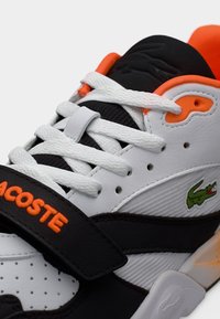 Baskets Lacoste blanches, noires et orange avec des lacets blancs, une sangle noire avec un texte orange et un logo crocodile vert sur le côté.