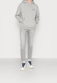 Sudadera gris con capucha y bolsillo delantero, combinada con vaqueros gris claro y zapatillas blancas con detalles azules y verdes.