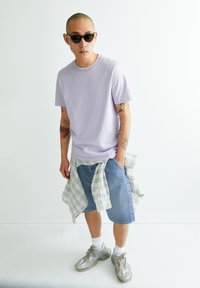 Hellblaues Baumwoll-T-Shirt in lockerer Passform, getragen mit blauen Denim-Shorts. Ein kariertes Hemd in Weiß und Blau um die Hüfte gebunden. Graue Turnschuhe.