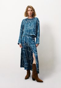 Türkisfarbiges zweiteiliges Outfit mit einem kastigen Langarmoberteil und einem Maxirock, der weiße abstrakte Linien aufweist, ergänzt durch braune Absatzstiefel.