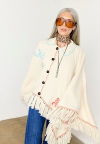 Γυναίκα που φοράει κρεμ πόντσο με φουντίτσες και κεντημένα πέταλα αλόγου και άλογο, μπλε τζιν, κασκόλ καφέ με μοτίβο παϊέτες και μεγάλα πορτοκαλί γυαλιά ηλίου.