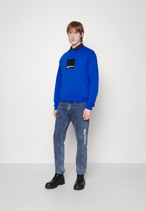 Felpa blu con toppa nera con logo, abbinata a jeans blu chiaro con scritte bianche e stivali neri stringati.