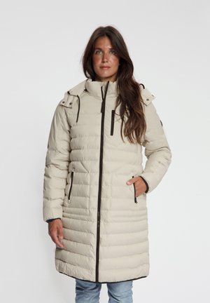 Vrouw draagt een lange beige doorgestikte winterjas met zwarte ritsdetails en capuchon, staand tegen een effen witte achtergrond.