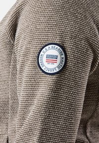 Giacca a righe marrone e crema con un design a maglia testurizzata, caratterizzata da una toppa circolare Seaside Nautical Legacy sulla manica.