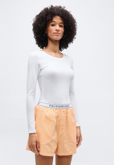 Γυναίκα με σγουρά μαλλιά που φοράει λευκή μπλούζα με μακριά μανίκια και πορτοκαλί ριγέ μποξεράκια με ζώνη Polo Ralph Lauren.