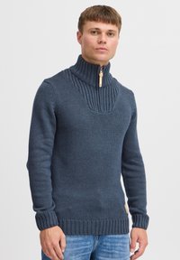 Dunkelblaues Strickpullover mit hohem Kragen, mit einem Rippmuster und einem halben Reißverschluss. Dehnbare Stoff mit strukturiertem Detail an den Schultern.