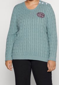 Ljusblå kabelstickad tröja med ribbade muddar och nederkant. Har tre knappdetaljer på axeln och broderad design på bröstet.