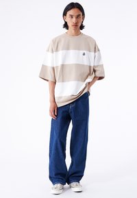 Beige und weiße gestreifte übergroße T-Shirt mit runder Ausschnitt, kombiniert mit locker sitzenden dunkelblauen Jeans und hellen Turnschuhen.