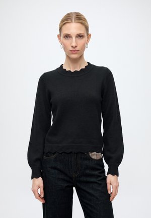 Femme aux cheveux blonds portant un pull noir à manches longues avec des bords festonnés, un jean sombre et de petites boucles d’oreilles créoles sur un fond uni.