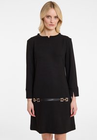 Schwarzes langärmliges Kleid mit Rundhalsausschnitt, ausgestattet mit goldenen Hardware-Details und einem Ledergürtel in der Taille. Glatte Textur, knielang.