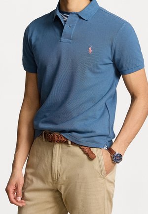 Mężczyzna ubrany w niebieską koszulkę polo z różowym logo, pasiastą podkoszulkę, beżowe spodnie, brązowy pleciony pasek oraz brązowy skórzany zegarek z niebieską tarczą.