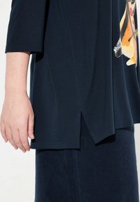 Topp marinblå laget av myk stoff med avrundet nederkant og slisser i sidene, med et dyremotiv på forsiden.