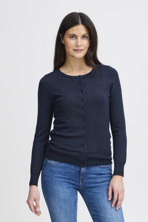 Ženska z dolgimi temnimi lasmi, oblečena v modro modem hlače s knoflji in modre kavbojke, stoji ob preprosti svetli podlagi.