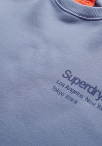 Sudadera azul clara hecha de tela suave. Presenta un cuello acanalado y texto impreso en azul oscuro que dice "Superdry" y nombres de ciudades.