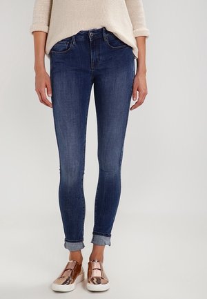 Partie inférieure du corps d'une personne portant un jean skinny bleu foncé retroussé, un pull beige et des baskets slip-on métalliques couleur or rose.