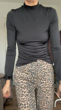 Črna dolga majica s turtleneck izrezom in oprijetim krojem, v kombinaciji s bež hlačami, ki imajo rjav leopardji vzorec in vidno gumbe.