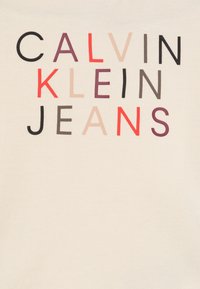 Krēmkrāsas kokvilnas T-krekls ar drosmīgu, daudzkrāsainu tekstu: "CALVIN KLEIN JEANS" dažādos lielajos burtiņos.