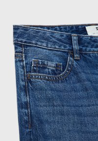 Detailní záběr modrých džín s přední kapsou, malou kapsou na mince a viditelným prošíváním poutek na opasek.