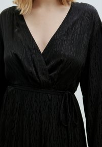 Schwarzes Wickelkleid mit strukturiertem Muster, tiefem V-Ausschnitt, langen Ärmeln und einem Bindegürtel in der Taille. Glattes Material mit dezenter glänzender Oberfläche.