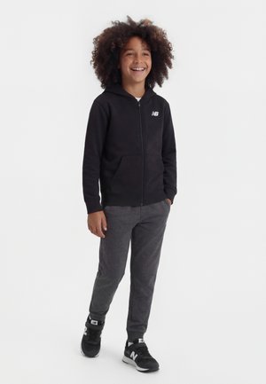 Bambino con capelli ricci in piedi e sorridente, che indossa una felpa con cappuccio nera con cerniera, pantaloni grigi e scarpe da ginnastica nere su uno sfondo bianco.