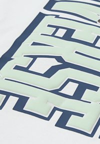 Hvitt bomullstoff har store, lagdelte tekster i lysegrønn og marineblå, med en teksturert finish og fet bokstavdesign.