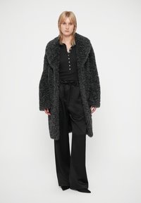 Blond kvinna som bär en lång, mörk krullig pälsrock över en svart knappad skjorta och vida svarta byxor, stående mot en enfärgad bakgrund.