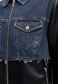 Κοντινή λήψη ενός μπουφάν με φθαρμένη μπλε denim πάνω περιοχή και λεία μαύρη δερμάτινη κάτω περιοχή, με μεταλλικά κουμπιά και φερμουάρ.