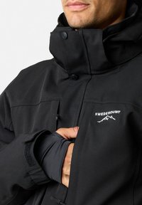 Zwarte softshell jack met hoge kraag, voor knopen en verstelbare manchetten. Voorzien van het witte "SWEDEMOUNT" logo en duurzaam materiaal.
