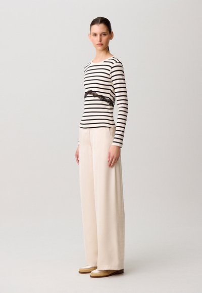 Femme debout portant un haut à manches longues rayé noir et blanc avec un détail en dentelle, un pantalon large crème et des chaussures beige.