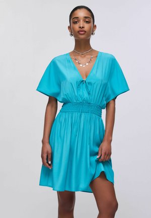 Žena u jarko plavoj haljini s V-izrezom, širokim rukavima, nabranim pojasom i slojevitim srebrnim ogrlicama na jednobojnoj svijetloj podlozi.